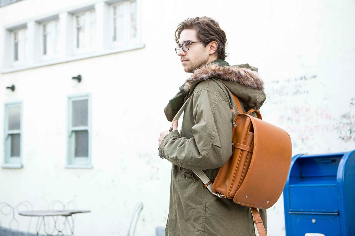 Man Wearing a Green Coat with Fur Hood and Brown Backpack Standing Outdoors.
